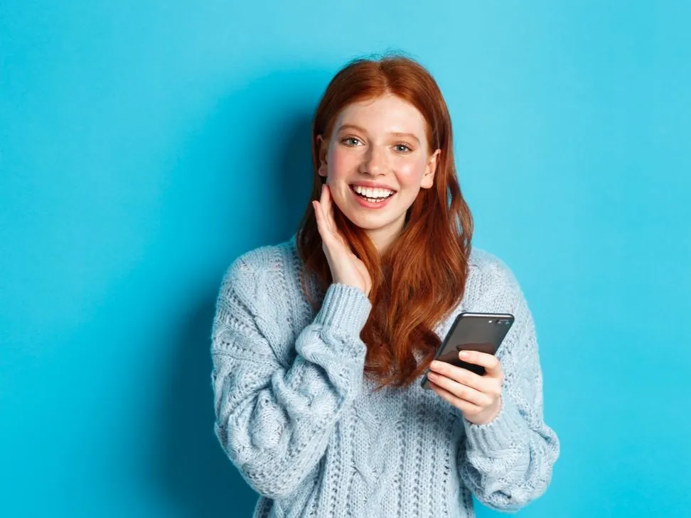 Beautiful redhead girl in sweater smiling at camera using mobile phone app standing with smartphone
